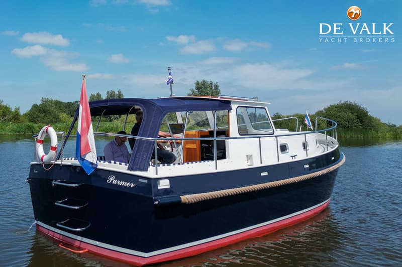 Een blauw-witte motorboot met rode onderkant en de naam "Purmer" vaart op een waterweg met groene oevers.