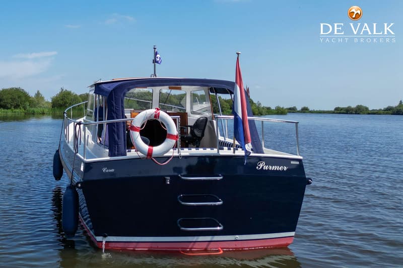 Een donkerblauwe boot met de namen "Cora" en "Purmer" op de spiegel, drijvend op water met een Nederlandse vlag.