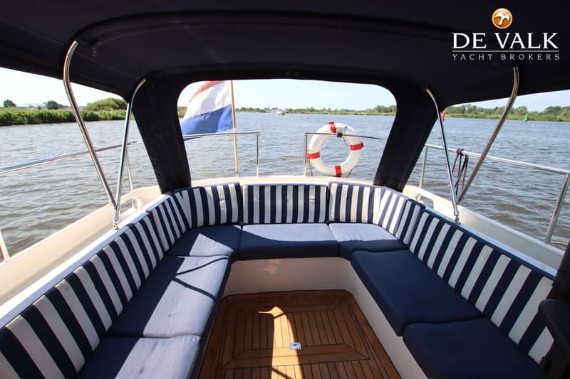 De foto toont het achterdek van een boot met blauw-wit gestreepte kussens en een houten vloer, uitkijkend over water.