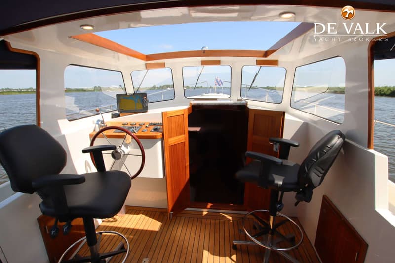 De foto toont de stuurhut van een boot met twee zwarte stoelen, een houten stuurwiel en een navigatiescherm.