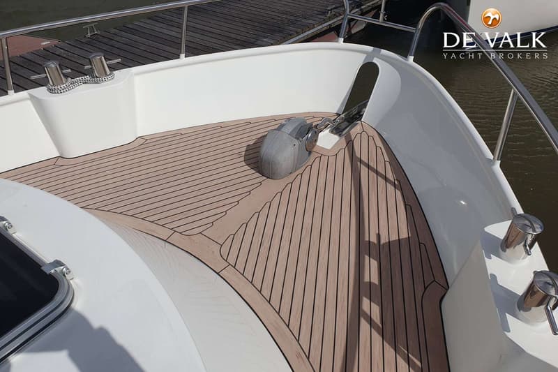 De foto toont het voordek van een boot met een teakdek, een ankerlier en twee bolders met een touw.