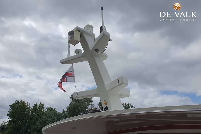 De foto toont een deel van de mast van een boot met navigatieverlichting, antennes en een vlag, tegen een bewolkte lucht.