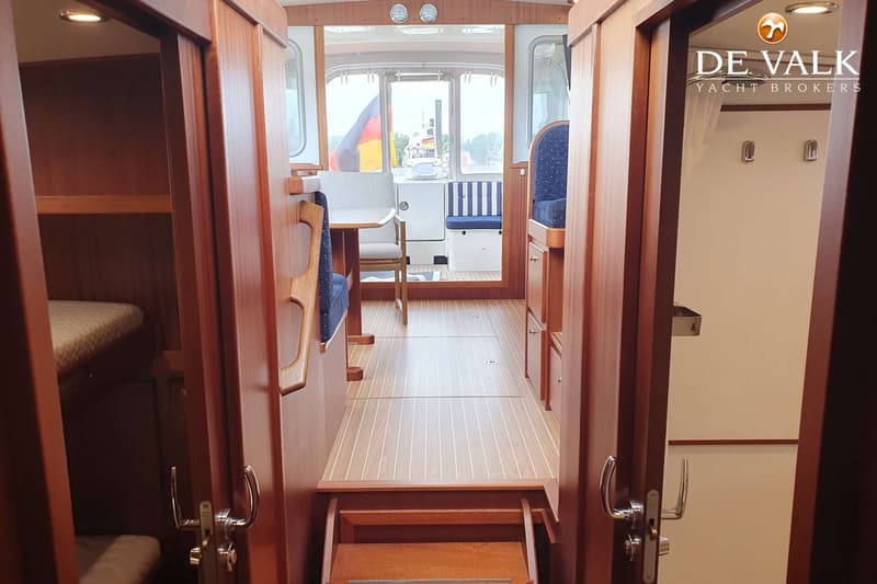 Een interieur van een boot met houten vloer en wanden, met een tafel en zitbanken, en een Duitse vlag zichtbaar door de…