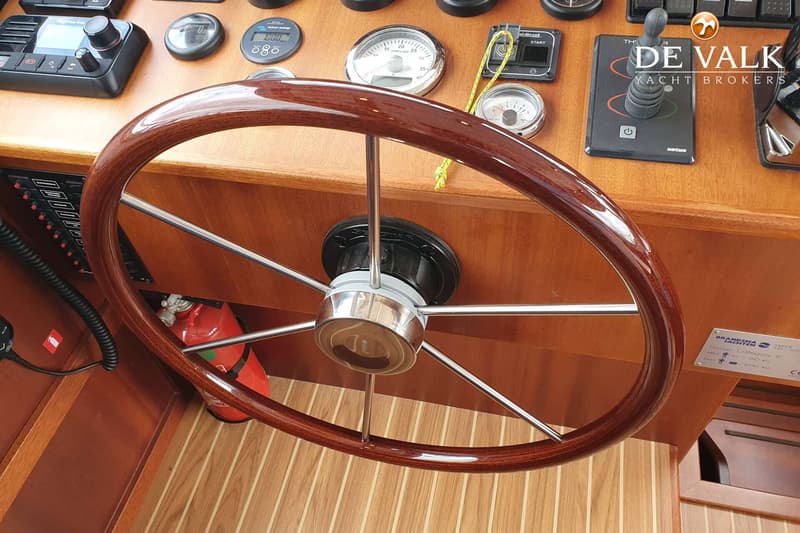 Een houten stuurwiel met chromen spaken is te zien, met daarachter een dashboard met diverse meters en schakelaars.