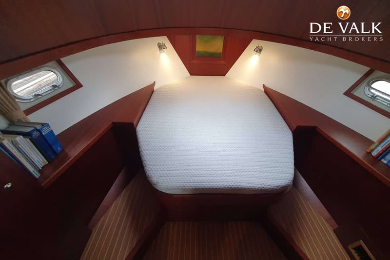 De foto toont een kajuit met een bed in de boeg, twee patrijspoorten, twee leeslampjes en boeken op planken.