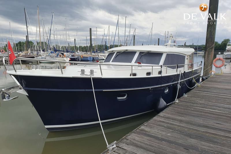 Een blauw-witte motorboot ligt afgemeerd aan een houten steiger, met zeilboten op de achtergrond onder een bewolkte lucht.
