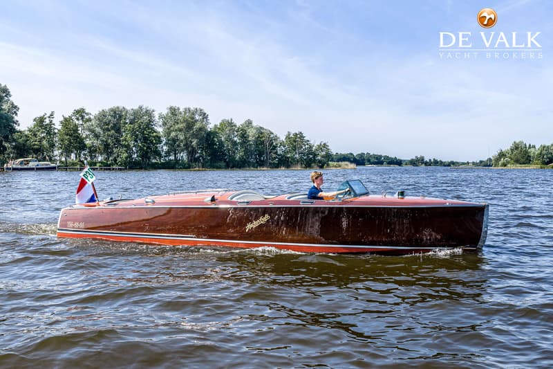 Een houten speedboot met een persoon aan het roer vaart op een waterweg met bomen op de achtergrond.