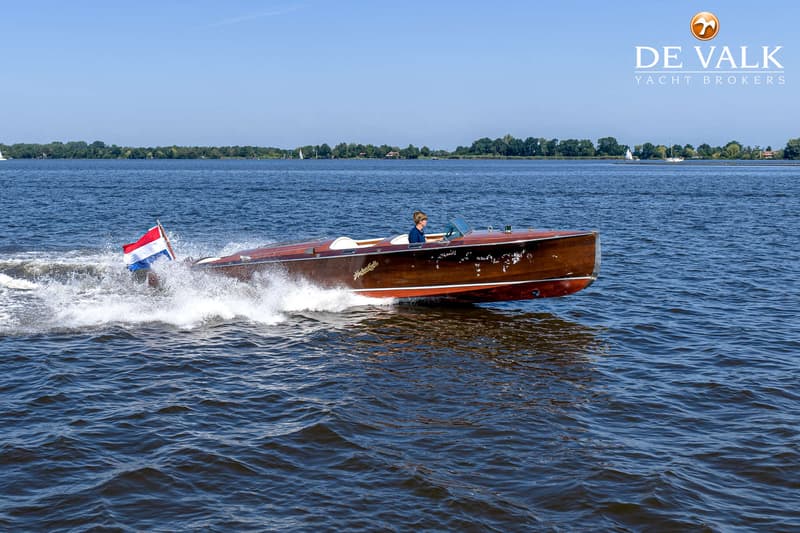 Een houten speedboot met een Nederlandse vlag vaart over het water, met een persoon aan het roer.