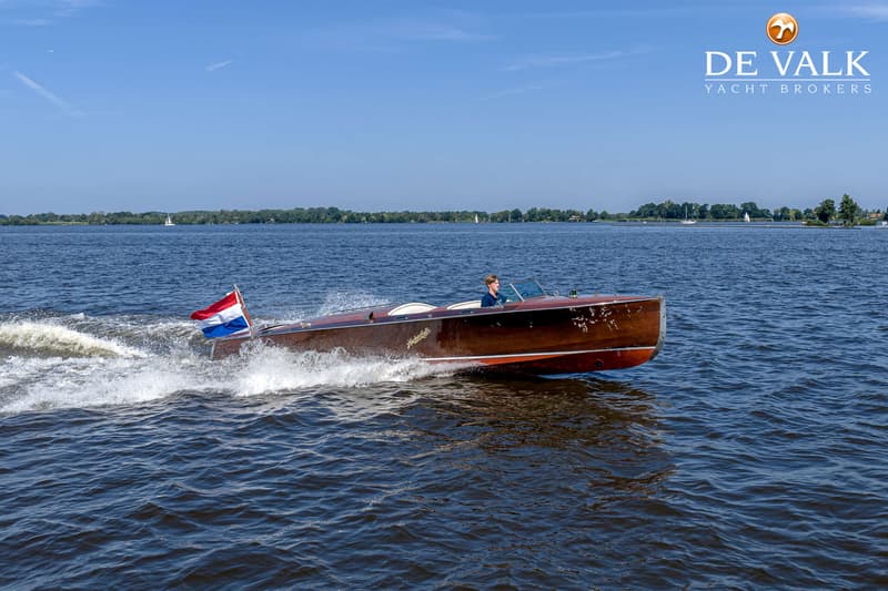 Een houten motorboot met een Nederlandse vlag vaart over het water, met een persoon aan het roer.