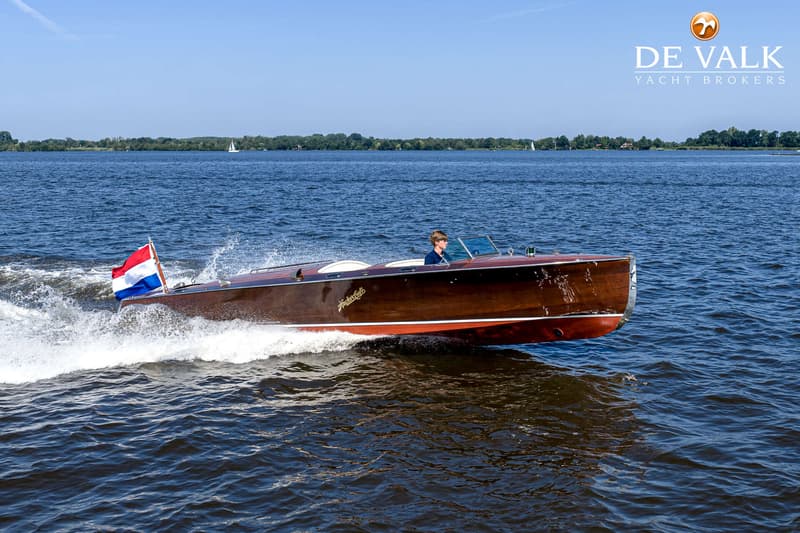 Een houten motorboot met een Nederlandse vlag vaart over het water, met een persoon aan het roer.