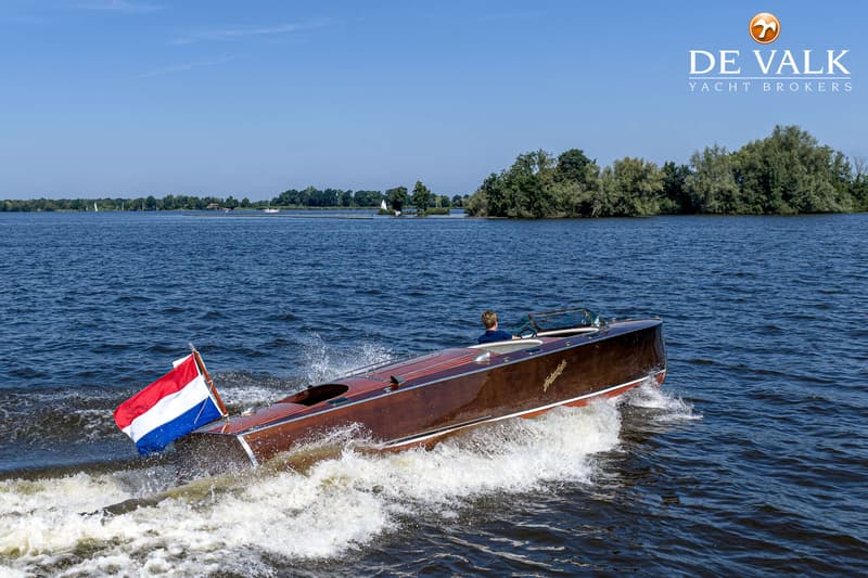 Een houten speedboot met een Nederlandse vlag vaart over het water, met een persoon aan het roer.
