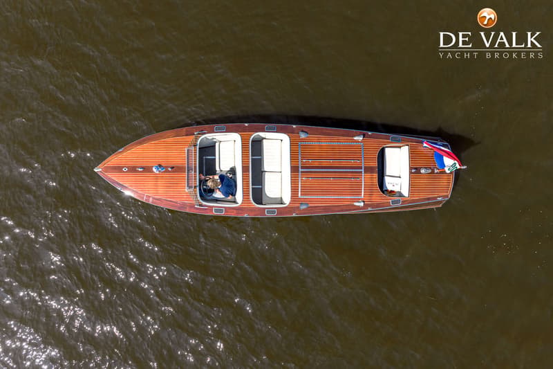 Een bovenaanzicht van een houten boot met een persoon aan het stuur, varend op donker water.