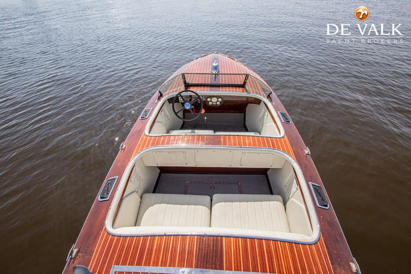 Een houten speedboot met een stuurwiel, dashboard en beige zitplaatsen, liggend in het water.