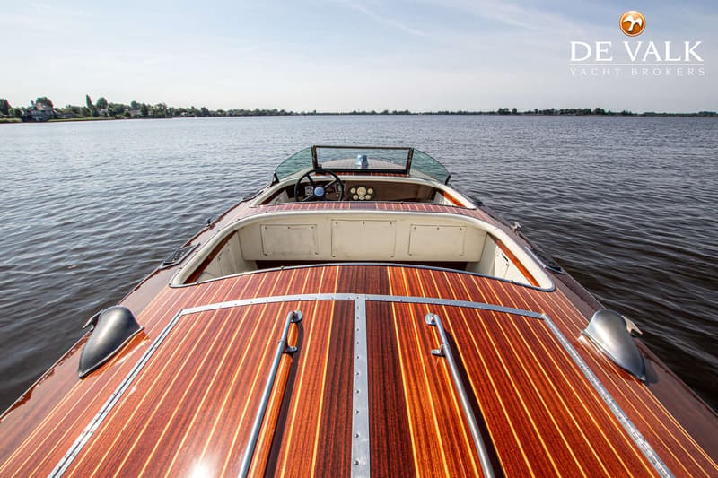 Een houten speedboot met een glanzende afwerking en een wit interieur vaart op een kalm meer onder een heldere hemel.