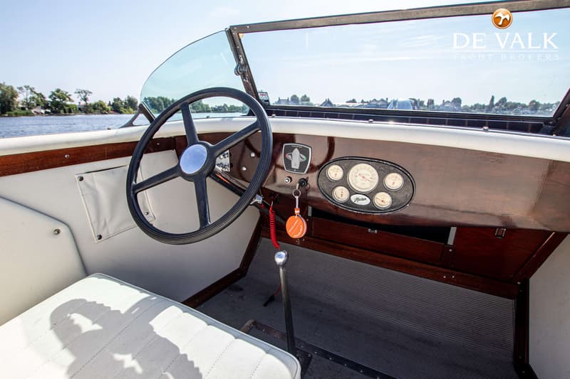 Een interieur van een boot met een stuur, een dashboard met meters, een versnellingspook en witte bekleding.