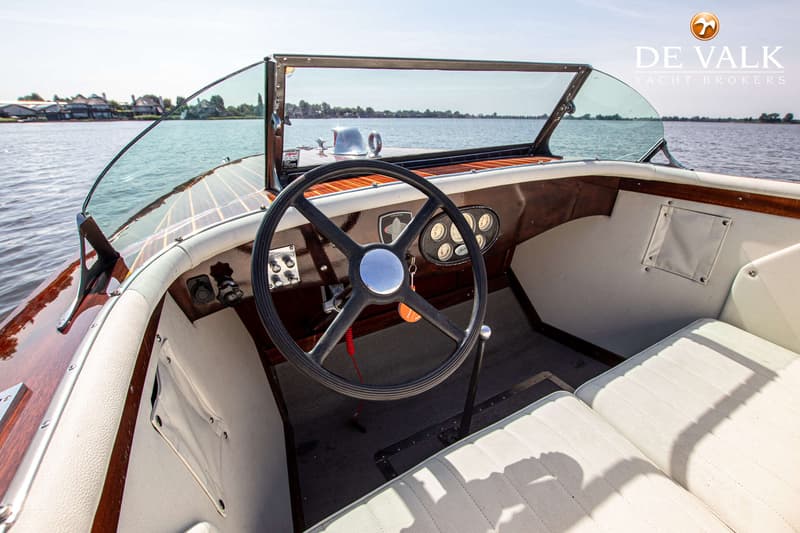Een interieur van een boot met een stuurwiel, dashboard met meters, witte bekleding en een houten afwerking.
