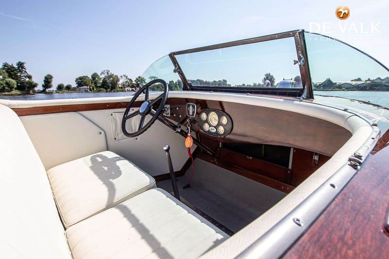 De foto toont het interieur van een boot met een stuurwiel, dashboard met meters, witte bekleding en een houten afwerking.