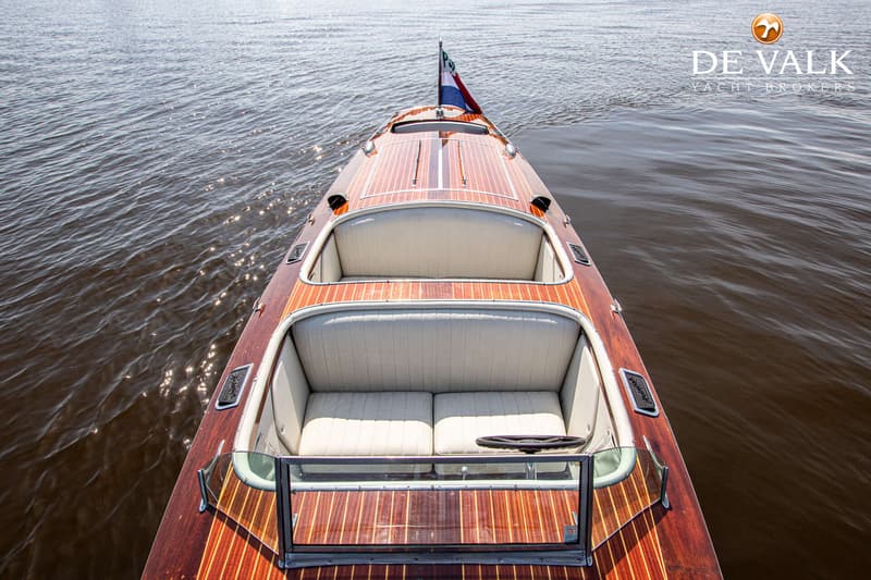 Een houten speedboot met twee zitgedeelten en een stuurwiel, varend op water met een Nederlandse vlag aan de boeg.