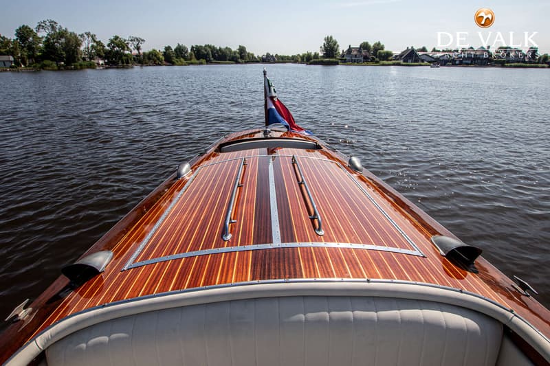 De voorkant van een houten boot met een Nederlandse vlag vaart over water met bebouwing op de achtergrond.