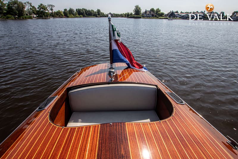 Een houten boot met een Nederlandse vlag op de boeg vaart op een waterweg met bebouwing en bomen op de achtergrond.