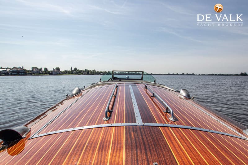 Een houten boot met een glimmend dek vaart op een meer met huizen aan de oever onder een blauwe lucht.
