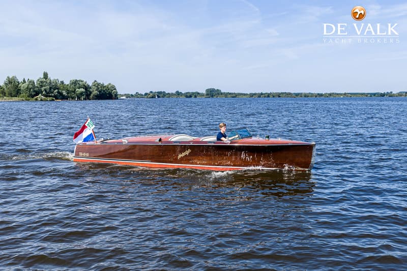 Een houten motorboot met een persoon aan het roer vaart op een waterweg met bomen op de achtergrond.