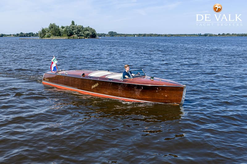 Een houten motorboot met een persoon aan het roer vaart op een meer met een eiland op de achtergrond.