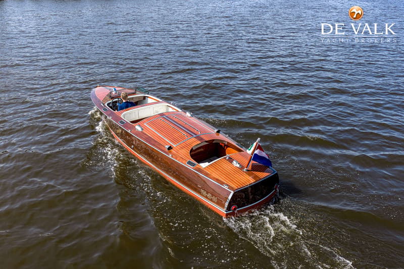Een houten motorboot met een Nederlandse vlag vaart op het water, met een persoon aan het roer.