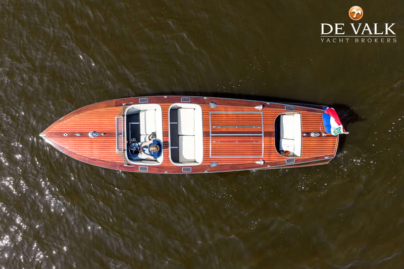Een houten speedboot met een persoon aan het stuur, drijvend op water, gezien van bovenaf.