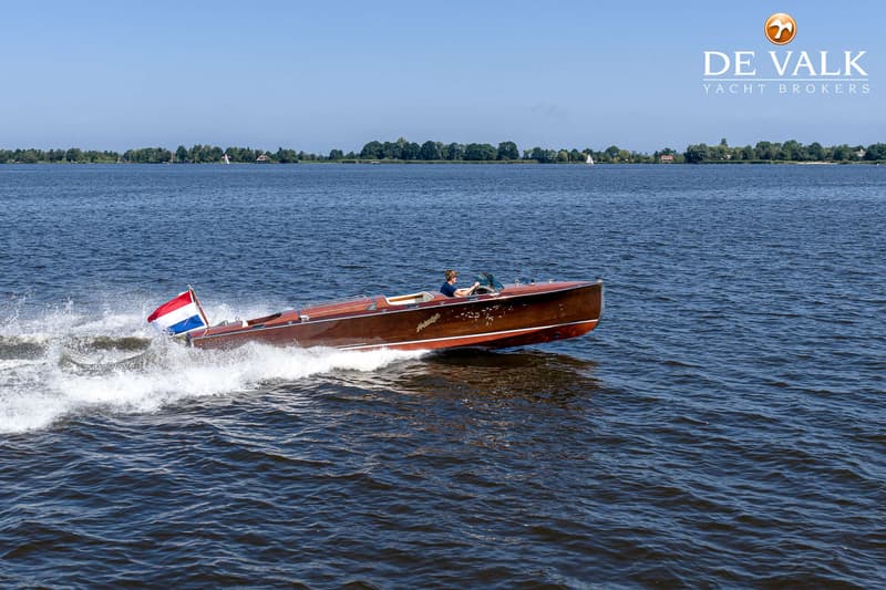 Een houten speedboot met een Nederlandse vlag vaart over het water, met een persoon aan het roer.
