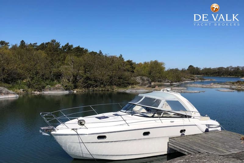 Een witte motorboot ligt afgemeerd aan een houten steiger, met een beboste kustlijn en rotsen op de achtergrond.