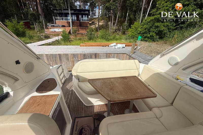 De foto toont het achterdek van een boot met crèmekleurige kussens, een houten tafel en een teakhouten vloer.