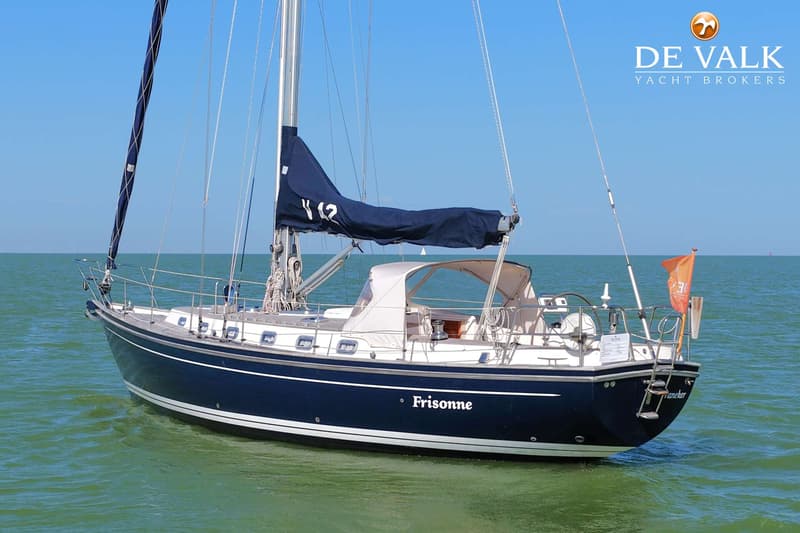 Een donkerblauwe zeilboot met witte opbouw en de naam "Frisonne" op de romp, ligt in groen water onder een blauwe lucht.