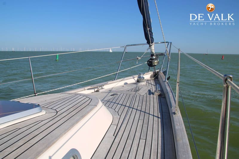 Een zeilboot met een opgerold zeil op de voorplecht, varend op open water met windmolens aan de horizon.