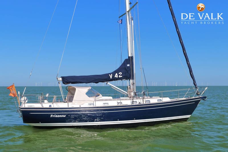 Een donkerblauwe zeilboot met de naam "Frisonne" en "V 42" op het zeil, drijft op groen water met windmolens op de…