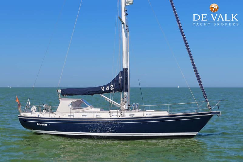 Een donkerblauwe zeilboot met de naam "Frisonne" en "V 42" op het zeil, drijft op groen water onder een helderblauwe lucht.