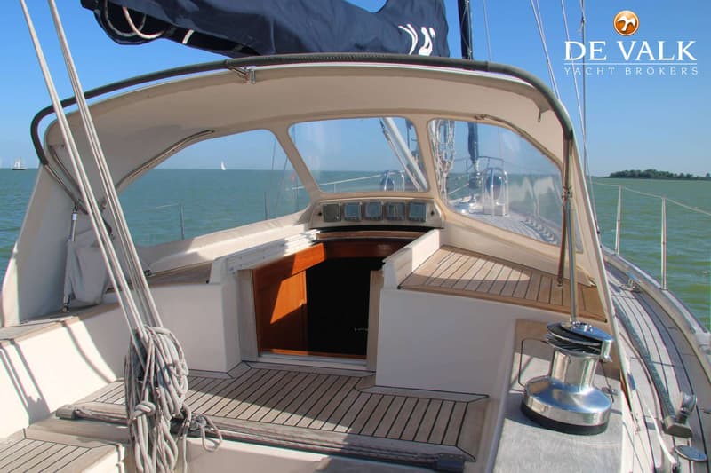 De foto toont het dek van een zeilboot met een open kajuitingang, een lier en touwen, onder een helderblauwe lucht.