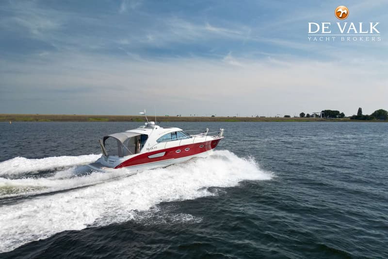 Een rood-witte motorboot vaart op het water en laat een wit schuimspoor achter, met een dijk op de achtergrond.