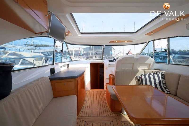 De foto toont het interieur van een boot met houten vloer, banken, een tafel, een tv en een stuurstand.