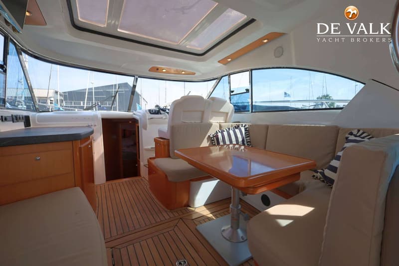 Een interieur van een boot met houten vloer, beige banken, een houten tafel en een stuurstand met witte stoelen.