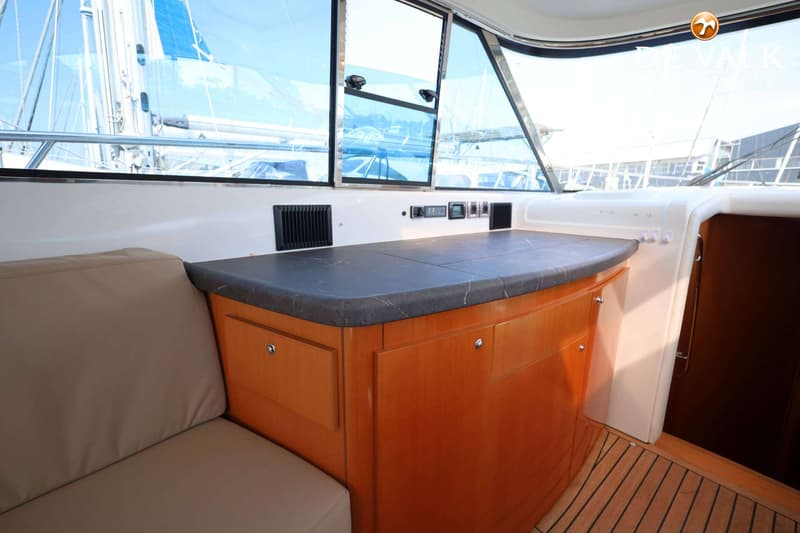 Een interieur van een boot met een beige bank, houten kasten, een donker aanrechtblad en ramen met uitzicht op een zeilboot.