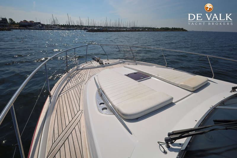 De foto toont het voordek van een boot met teakhouten afwerking, witte kussens en een reling, met een jachthaven op de…