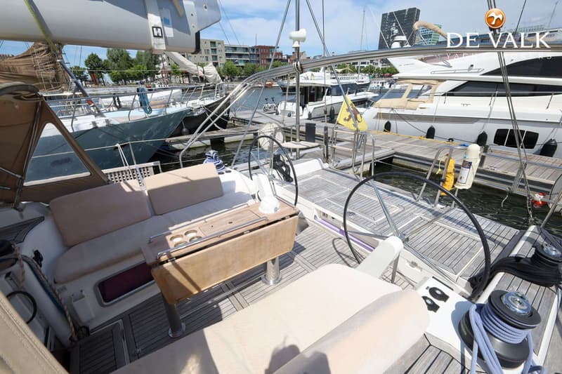 Een bovenaanzicht van het dek van een zeilboot met twee stuurwielen, een houten tafel en beige kussens.
