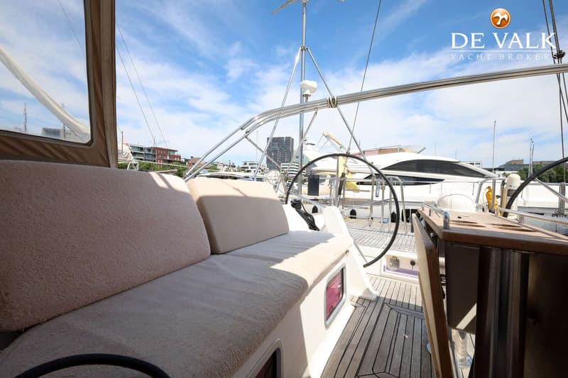 De foto toont het dek van een zeilboot met beige kussens, een stuurwiel en een houten tafel, onder een blauwe lucht.