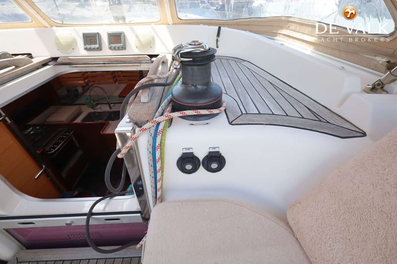 Een deel van de kuip van een zeilboot met een lier, touwen, een houten dek en de ingang naar de kajuit.