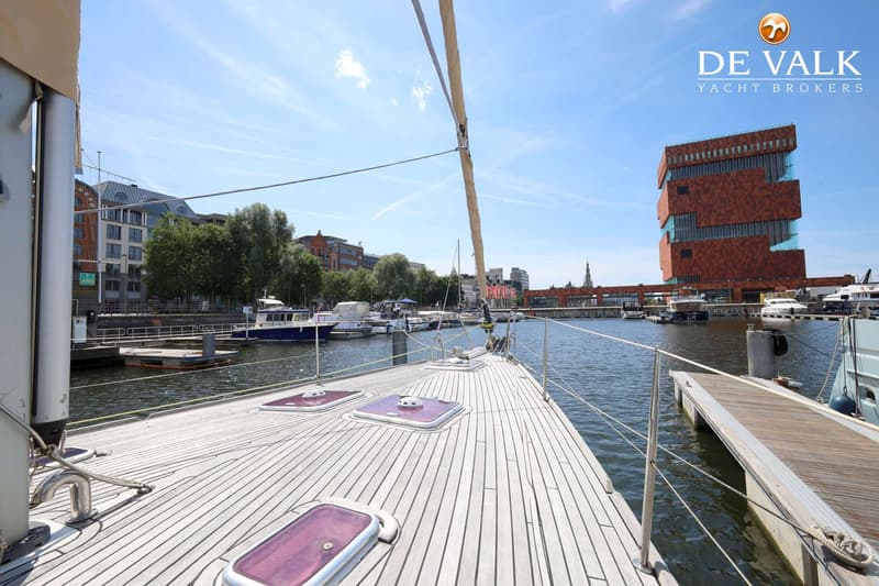 De foto toont het teakhouten dek van een zeilboot met drie luiken, liggend in een haven met gebouwen op de achtergrond.