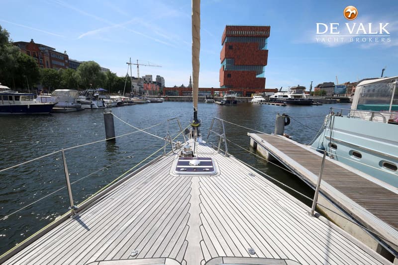 De foto toont het voordek van een zeilboot met een houten dek, een mast en een ankerlier, met een haven op de achtergrond.