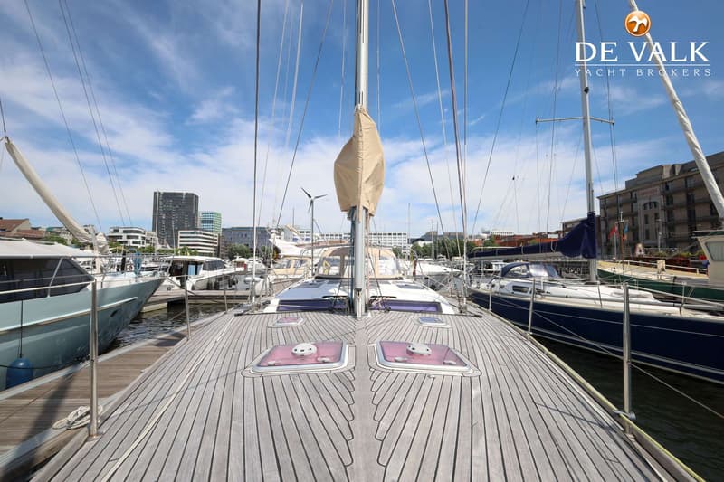 Een teakdek met twee luiken en een mast met opgerold zeil, omringd door andere boten in een haven.