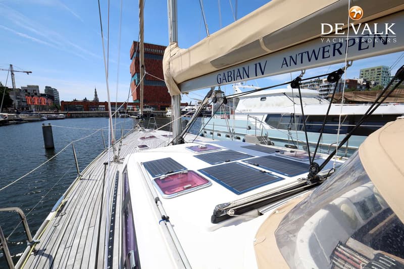 Een zeilboot met een houten dek en zonnepanelen op het dak, aangemeerd in een haven met gebouwen op de achtergrond.