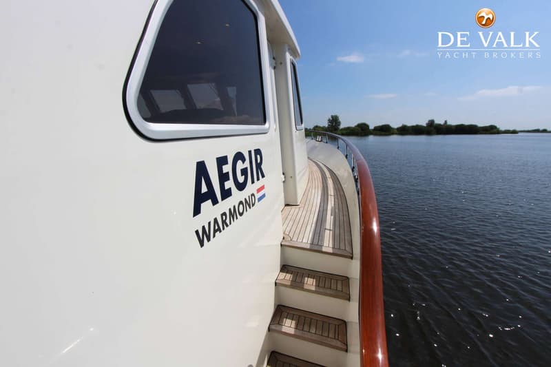 Een witte boot met de naam "AEGIR WARMOND" en de Nederlandse vlag, met een houten dek en trap, vaart op water.